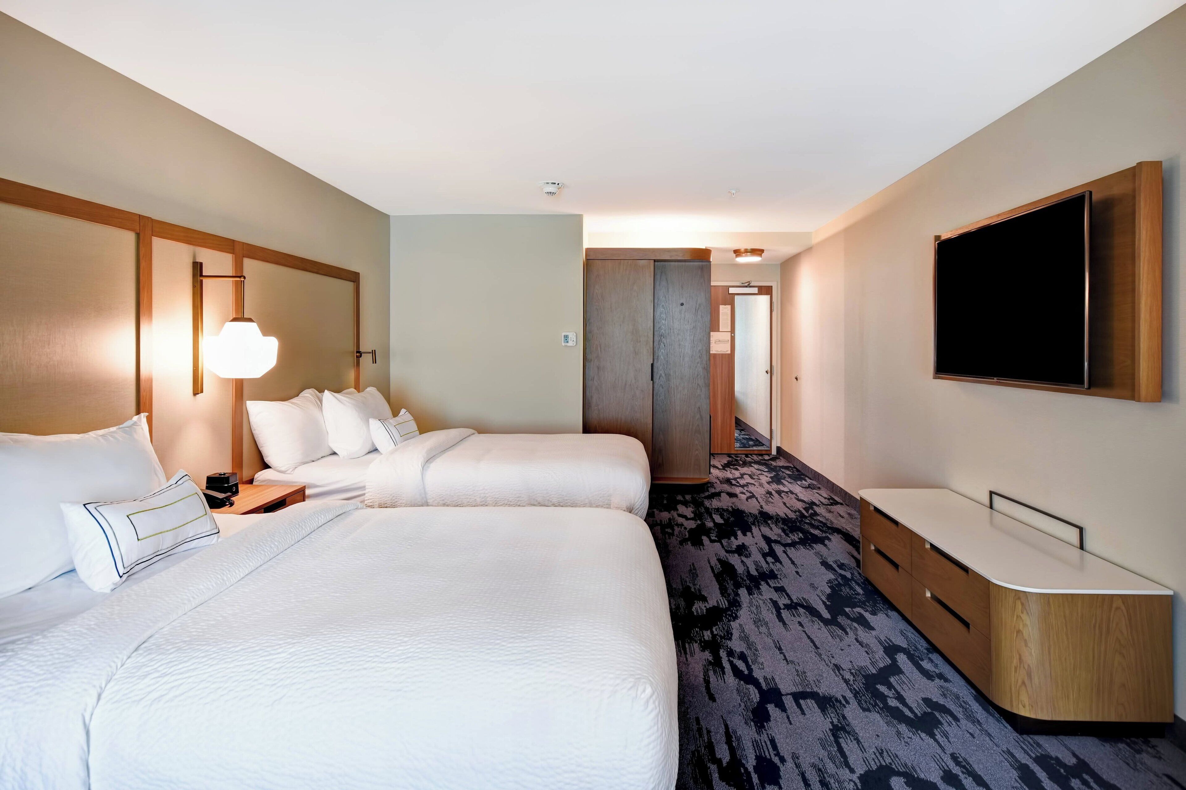 A hotel room with two beds, a desk, a flat-screen TV, and a carpeted floor.