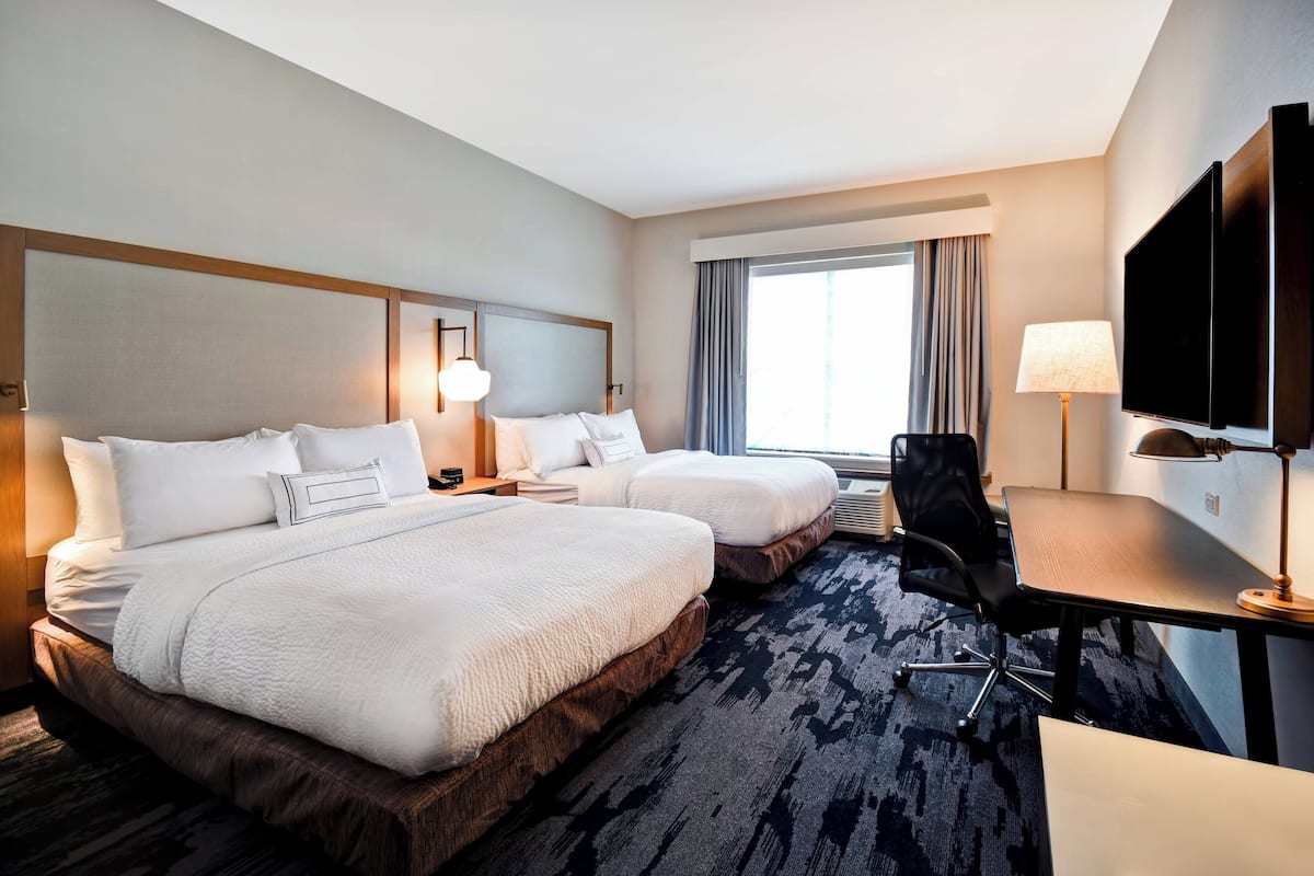 A hotel room with two beds, a desk, a chair, a TV, and a large window with curtains.