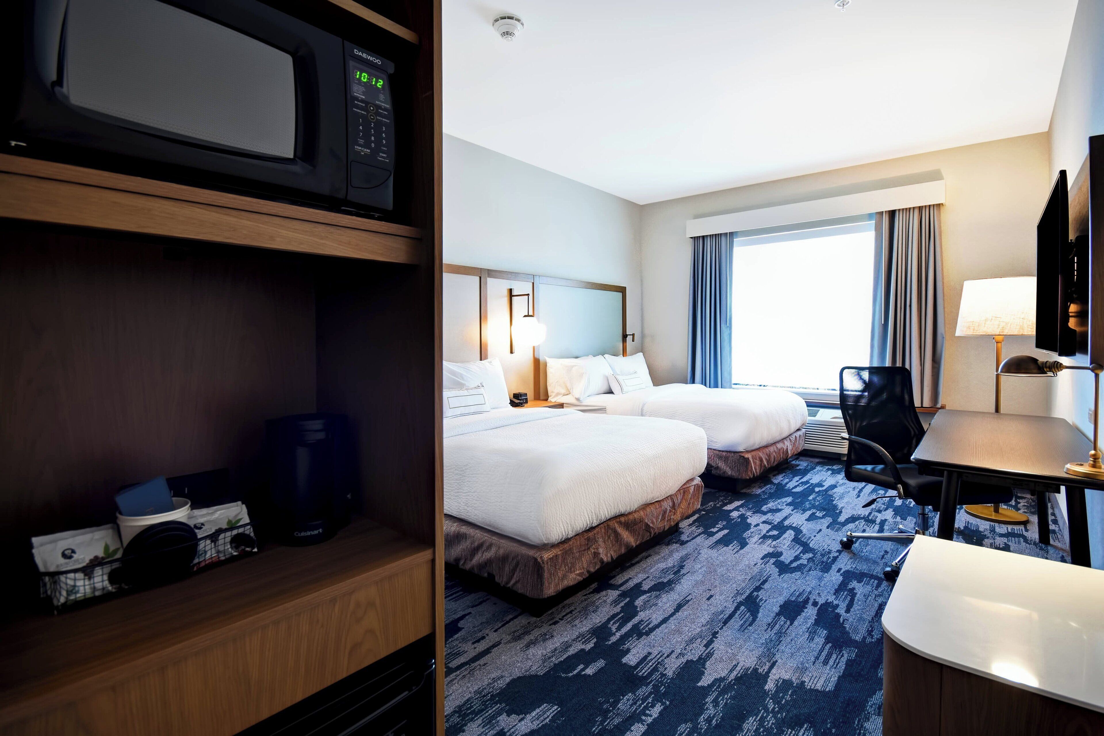 A hotel room with two beds, a desk, a chair, a microwave, and a large window with curtains.