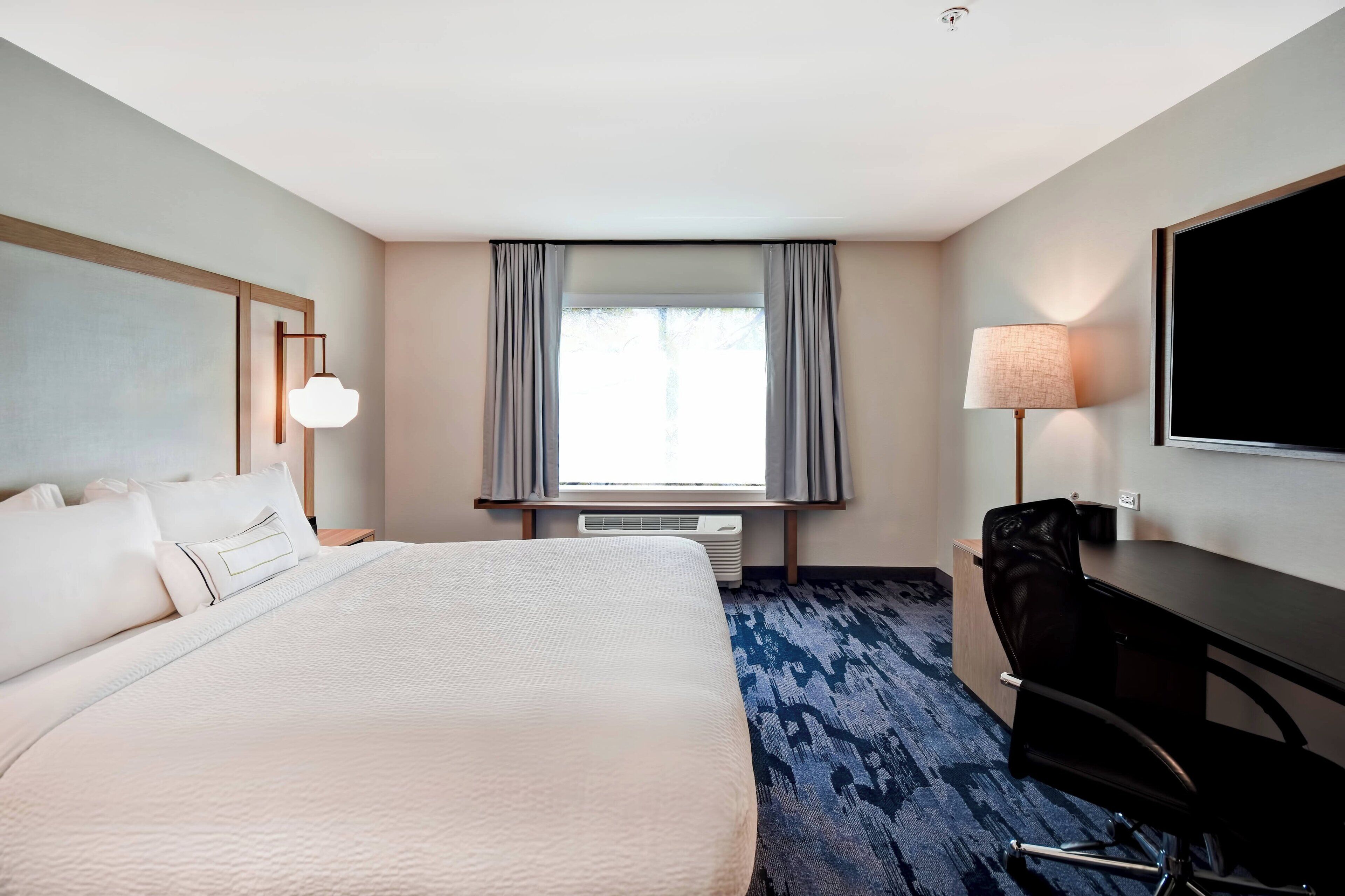 A hotel room with a large bed, a desk, a chair, a TV, and a window with curtains.