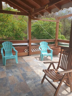 A covered outdoor seating area with two turquoise chairs and one wooden chair, surrounded by a wooden fence and potted plants.