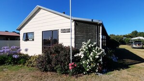Un piccolo edificio bianco con tetto spiovente, una finestra e una porta. Davanti all'edificio si estende un giardino con fiori viola e bianchi.