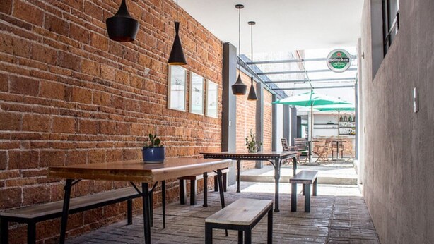 An outdoor seating area with brick walls, wooden tables, benches, hanging pendant lights, and a view of the street.