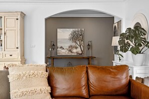 Uma sala de estar com um sofá de couro marrom, uma poltrona bege, um aparador de madeira, uma pintura emoldurada de uma praia e um vaso com planta.