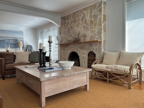 Uma sala de estar com lareira de pedra, um sofá de couro marrom, uma poltrona de vime e uma mesa de centro de madeira.