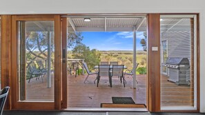 A sliding glass door leading to an outdoor deck with a table and chairs, a barbecue grill, and a view of a rural landscape. - Bungala Cottage - 28 Jetty Road (Normanville)
