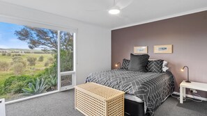 Ein Schlafzimmer mit einem großen Fenster, einem Bett mit gemusterten Bettwaren, einem Nachttisch und einem kleinen Holzhocker.
