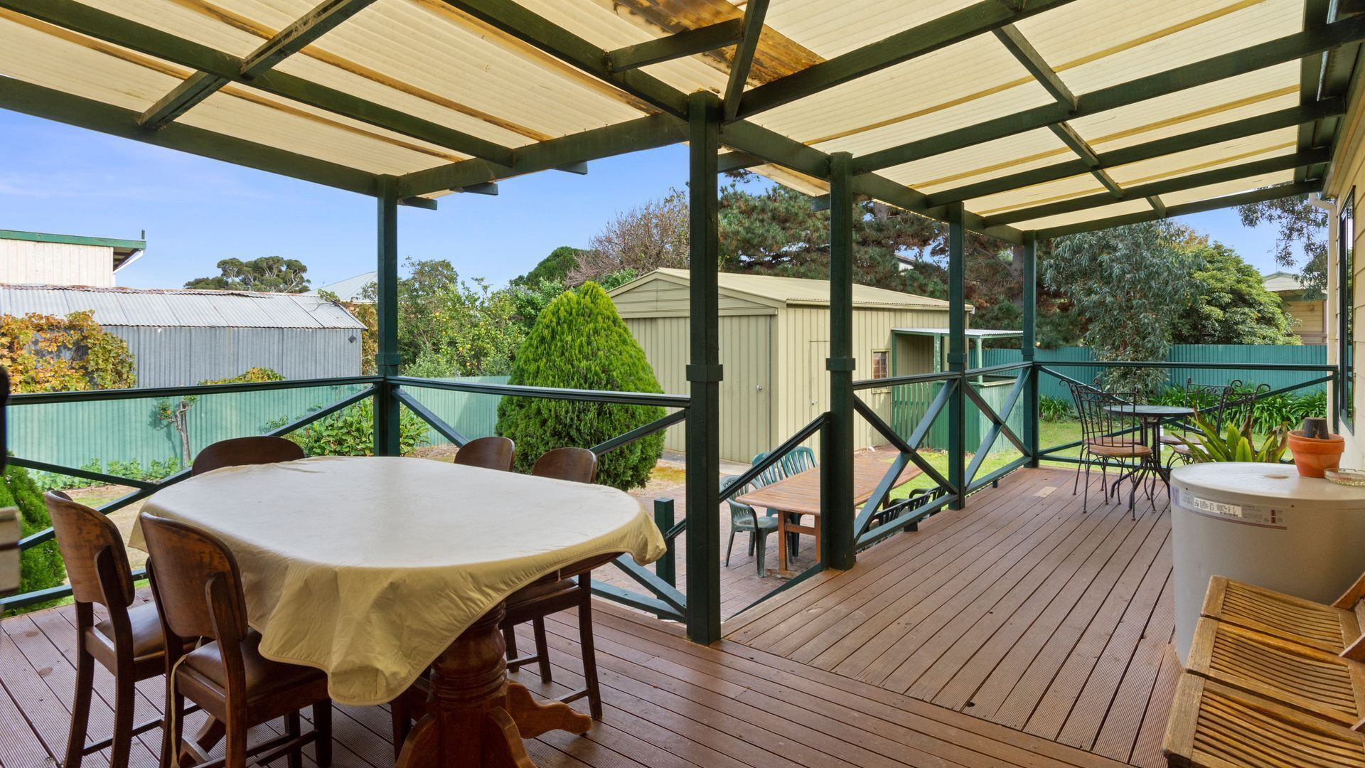 An outdoor covered patio with a table and chairs, surrounded by a wooden deck and a green fence.
