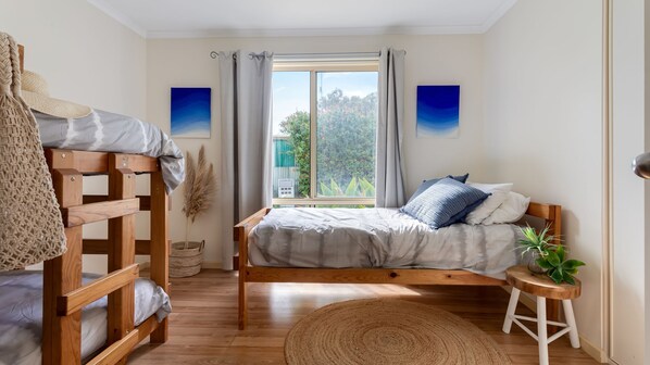 Un dormitorio con una cama litera, una cama individual, una ventana con cortinas, vistas a zonas verdes y una planta pequeña sobre un taburete.