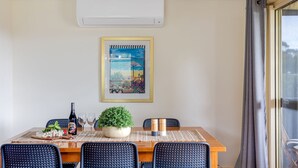 Een eetruimte met een tafel gedekt voor twee personen, een potplant en een lijst met een kunstwerk erboven.