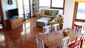 A spacious living area with a wooden floor, a sofa set, a television, and a dining table with chairs. - Tree Top - 4 Robert Norman Street (NORMANVILLE)