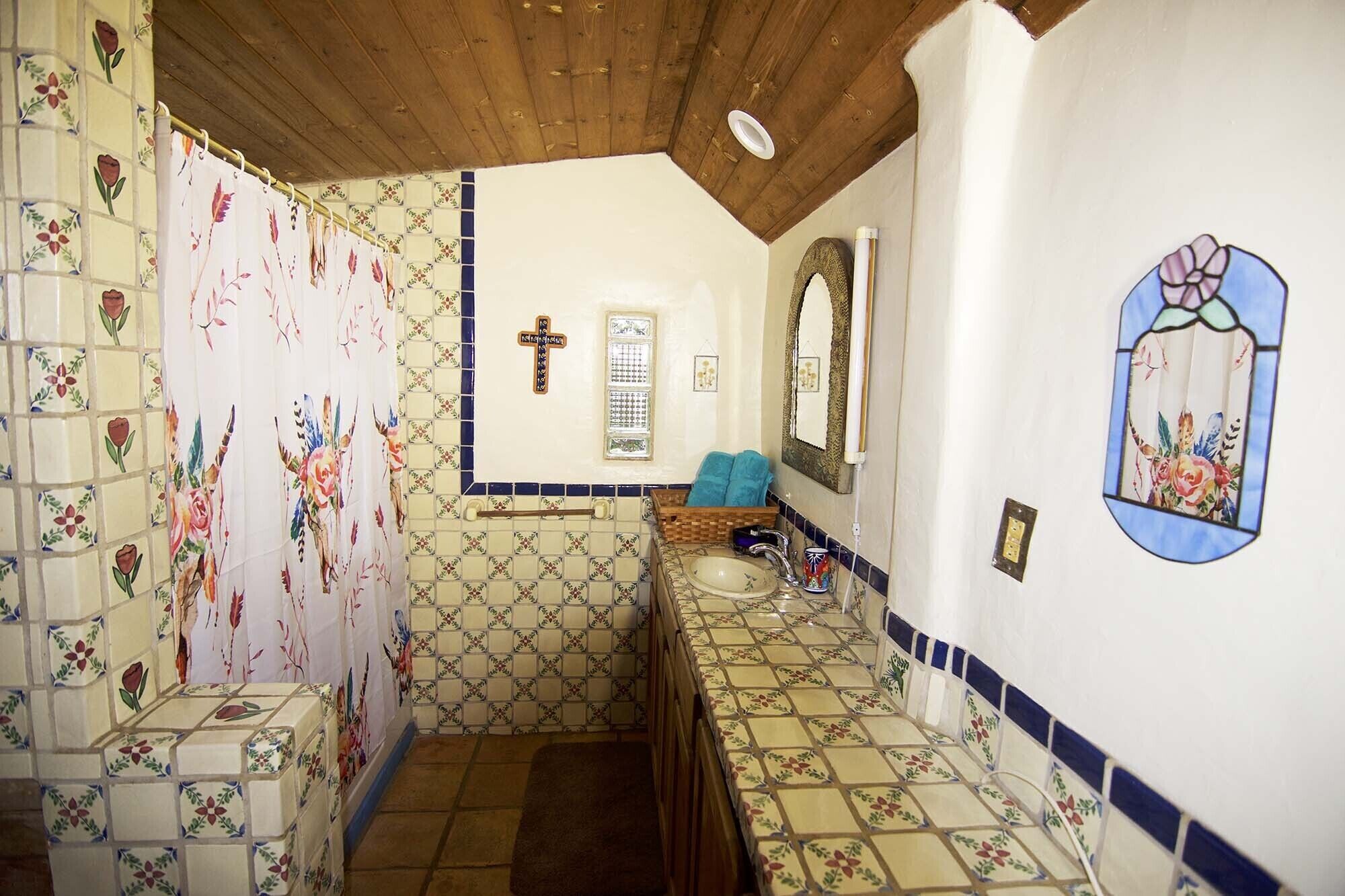 Un baño con patrones de azulejos florales, techo de madera, lavamanos con espejo y una cruz en la pared.