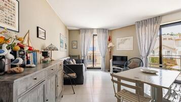 A modern living room with a sofa, dining table, and a TV. There is a balcony with curtains and a view of the outside.