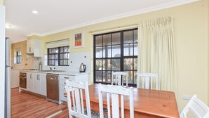 A kitchen with white cabinets, a wooden dining table, and a large window with curtains. - By the Beach - 30 Jetty Road (Normanville)