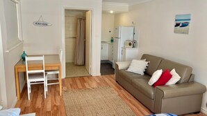 A living room with a beige sofa, a small table, and a chair. There is a kitchen area visible through an open door. - By the Beach - 30 Jetty Road (Normanville)