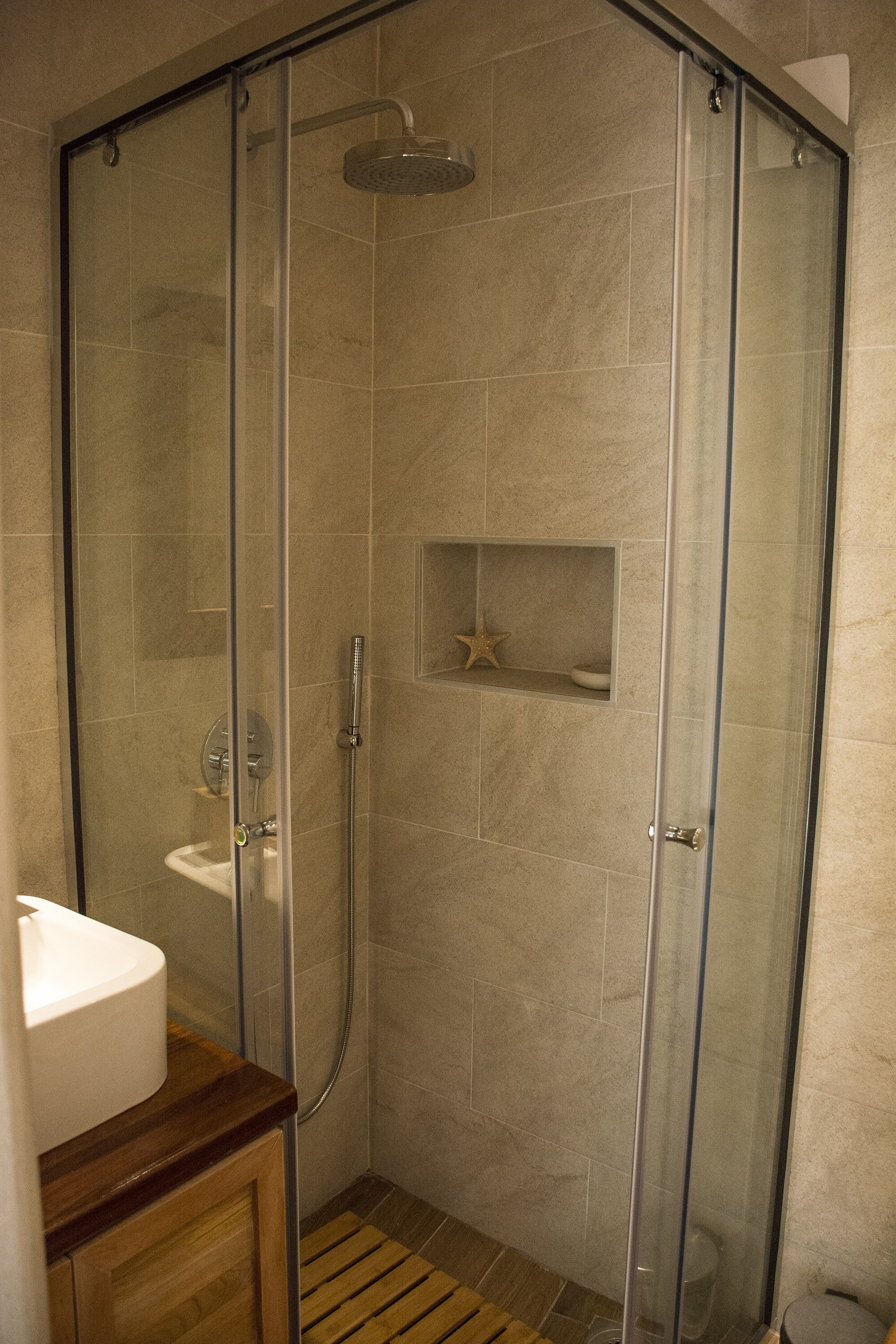 A glass-enclosed shower with a handheld showerhead, a built-in shelf with a starfish decoration, and a wooden vanity with a sink.