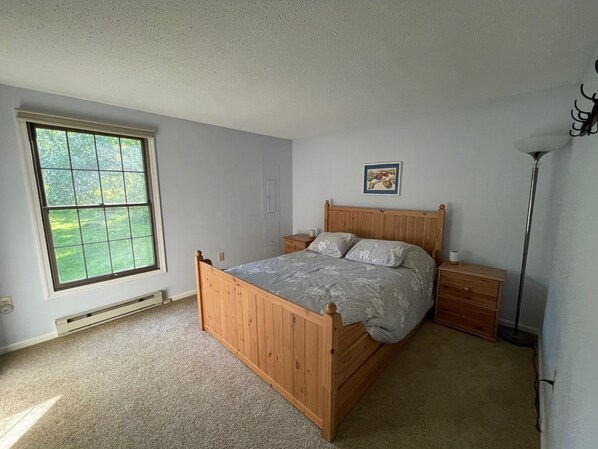 A bedroom with a wooden bed, a nightstand, a bedside table, a floor lamp, and a window with a view of greenery.