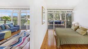 Relax in this serene bedroom with a large bed, sofa, and sliding glass door leading to an outdoor deck with a table and chairs overlooking a lush landscape.
