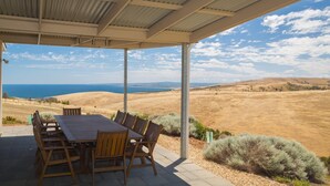 Un espacio de comedor al aire libre con techo, con una mesa y sillas, que ofrece vistas a un paisaje pintoresco con colinas y una masa de agua.