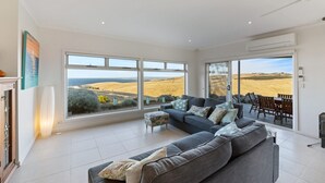 Un salón espacioso con un sofá grande, un comedor con sillas y vistas a un campo y al océano a través de amplias ventanas.