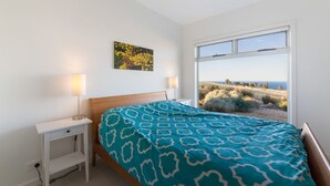 Un dormitorio con cama, mesita de noche, lámpara, un cuadro y ventana con vista al campo.