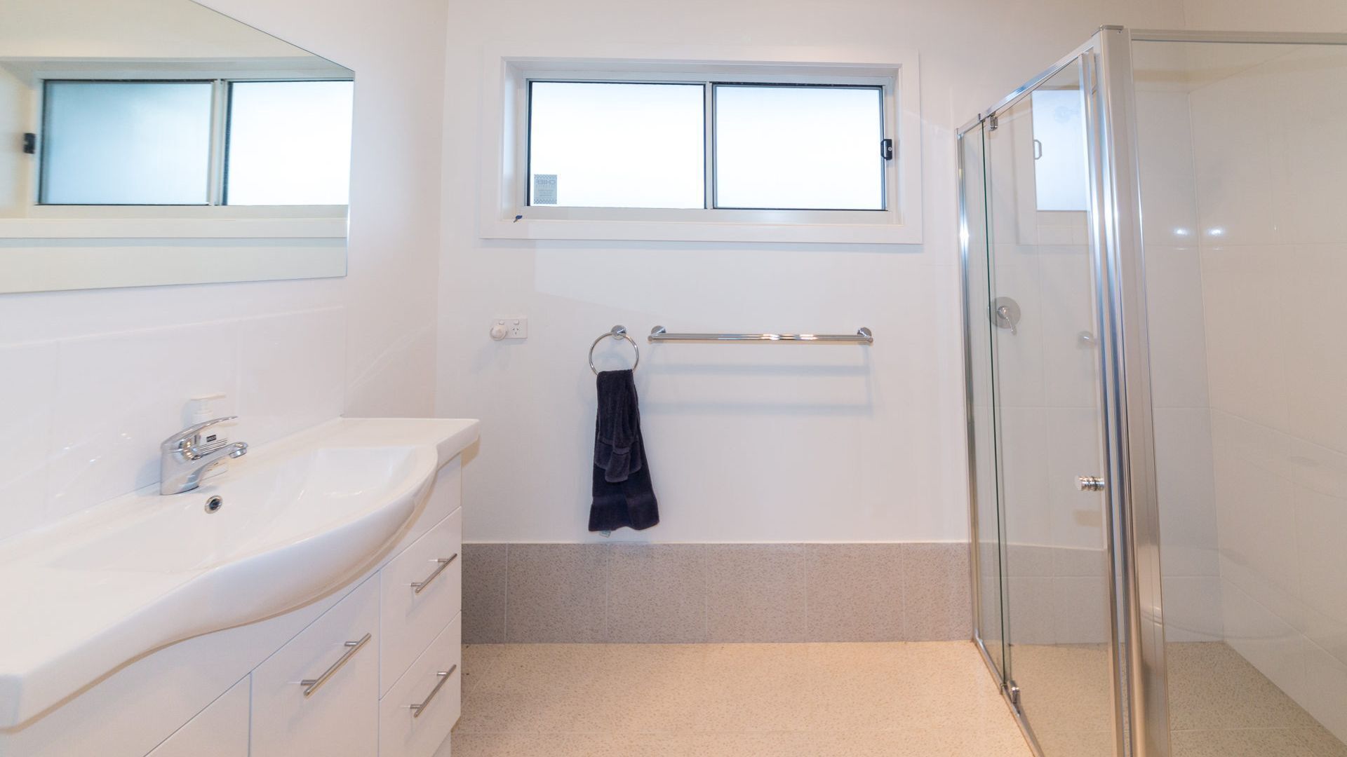 Une salle de bain avec un lavabo blanc, un miroir, un porte-serviettes, une fenêtre et une cabine de douche en verre.