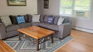 Olohuoneessa on harmaa kulmasohva, puinen sohvapöytä, kuviollisia mattoja ja taideteoksia seinillä.