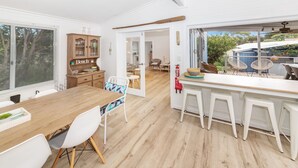 A modern kitchen with a wooden table and white bar stools, a wooden cabinet with glass doors, and a view of the outdoors. - Linga Longa - 20 Laronde (CARRICKALINGA)