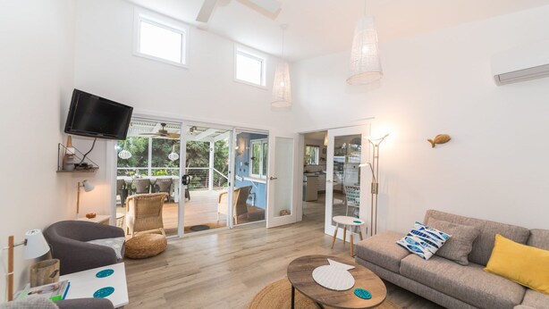 A modern living room with a grey sofa, a coffee table, and a TV mounted on the wall. There is a balcony with wicker furniture and a view of greenery. - Linga Longa - 20 Laronde (CARRICKALINGA)