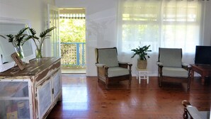 A room with wooden flooring, two armchairs, a small table, a television, and a window with sheer curtains. - Sawtells Cozy cottage (Sawtell)