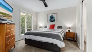 Une chambre avec un grand lit, une commode en bois, une table de chevet, un ventilateur de plafond et une fenêtre donnant sur une plage.