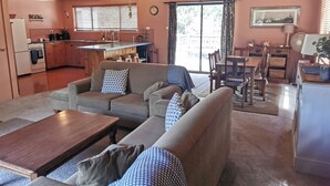 Un salon chaleureux avec un ensemble de canapés, une table basse en bois et un coin repas comprenant une table et des chaises. On aperçoit une cuisine équipée d’une cuisinière et d’un évier au fond.