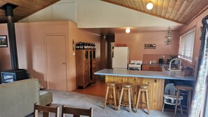Un salon chaleureux avec un plafond en bois, un canapé, une table à manger avec des chaises, une cuisine équipée d’un réfrigérateur, d’une cuisinière et de tabourets de bar.
