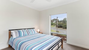 A bedroom with a bed, a nightstand, a lamp, and a large window with a view of a road and greenery. - Carrickalinga Shores - 35 Solitude Drive (CARRICKALINGA)
