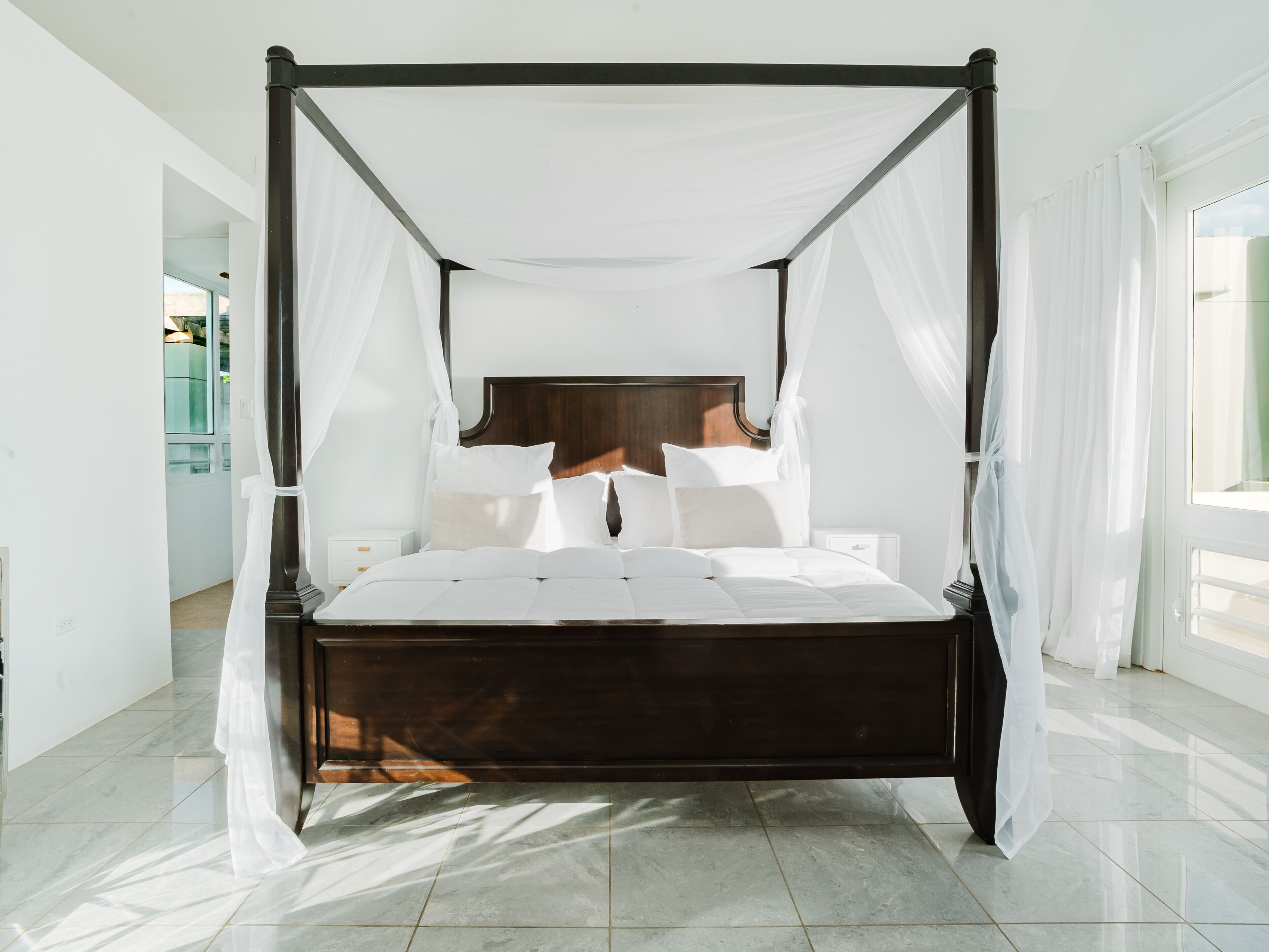 A four-poster canopy bed with white linens, a wooden headboard, and a marble floor.