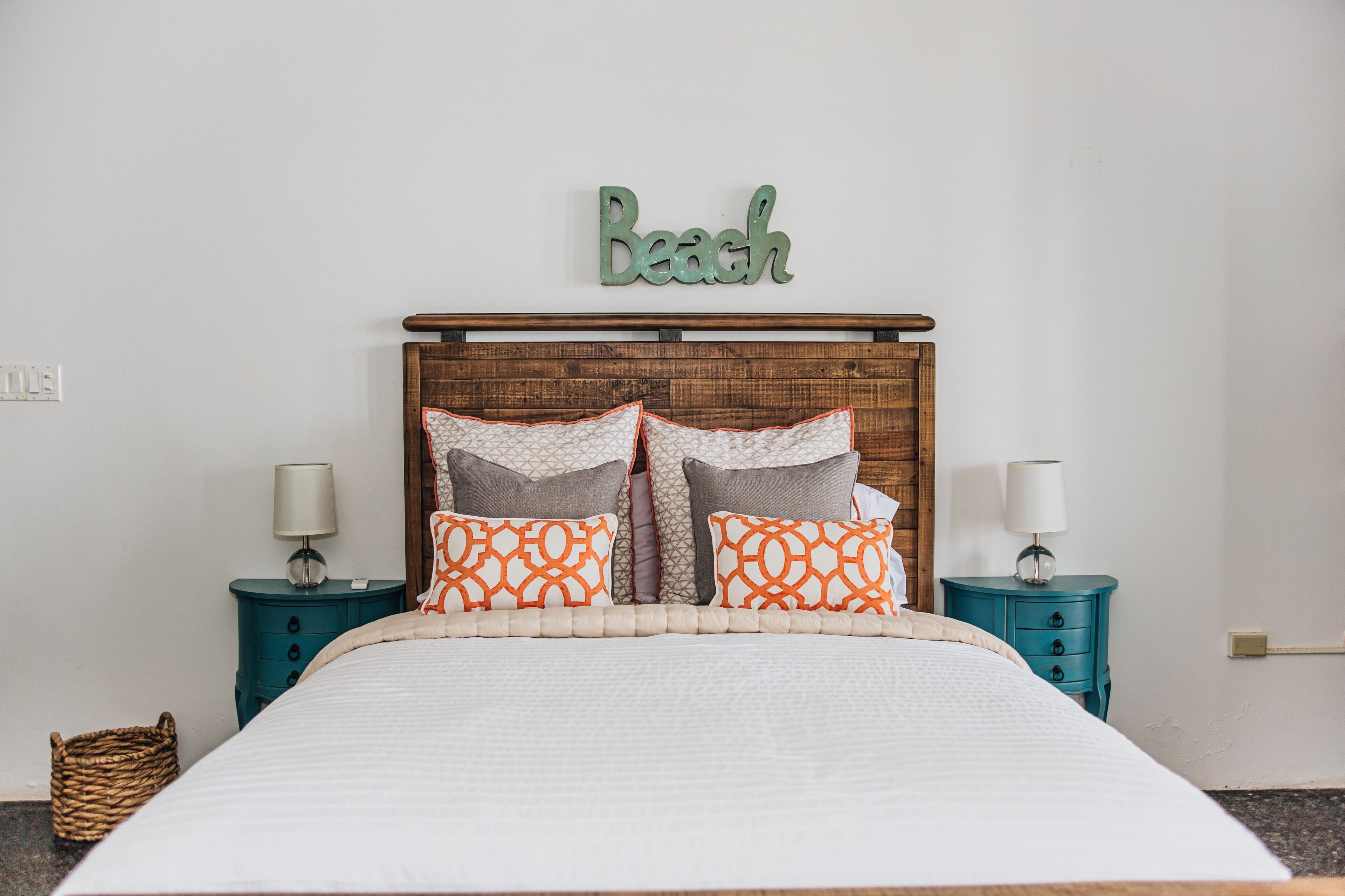 A neatly made bed with a wooden headboard, two matching nightstands, and a wall-mounted sign that reads "Beach".