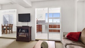 A living room with a television, a bookshelf, a dining area with a table and chairs, and a sliding glass door leading to a balcony. - Apartments at the beach (Blackmans Bay)