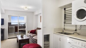A compact kitchen with a built-in washing machine, a dining area with a red ottoman, and a view of the outside through a glass door. - Apartments at the beach (Blackmans Bay)