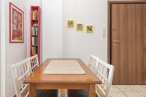 A dining area with a wooden table, white chairs, a woven placemat, and a red bookshelf with books. There are three framed pictures on the wall and a wooden door with a brass handle.