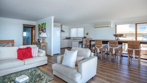 Uma sala de estar moderna com sofá, poltrona e mesa de centro. É visível uma área de jantar com cadeiras de vime e uma mesa. A cozinha fica ao fundo, com armários brancos e uma janela.