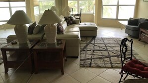A spacious living room with a sectional sofa, two lamps on side tables, a red chair, and large windows with blinds. - First Lido - New pool deck, and landscaping, 
New elevator, Freshly painted (Sarasota)
