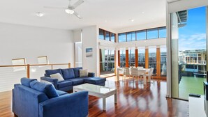 A spacious living area with a blue sectional sofa, a glass coffee table, and a dining table with chairs. Large windows offer views of a building and a body of water.