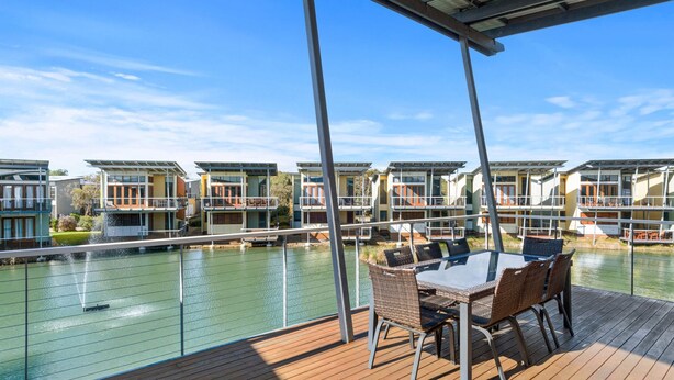 A deck with a table and chairs overlooking a body of water and a row of modern buildings.