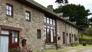 A stone building with multiple windows and a door, surrounded by greenery and a gravel path.