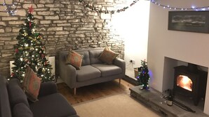 A cozy living room with a stone wall, a fireplace, a small Christmas tree, and a grey sofa.