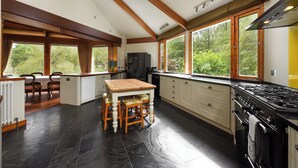 Une cuisine spacieuse dotée d’un grand îlot central, de poutres apparentes en bois au plafond et de grandes fenêtres offrant une vue sur l’extérieur.