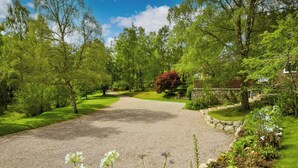 Une allée de gravier mène à une maison entourée d’une végétation luxuriante et de plantes en fleurs.