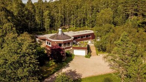 Une grande maison de plain-pied dotée d’une tour circulaire, entourée d’une forêt dense.