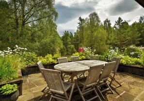 An outdoor patio with a round wooden table and six chairs, surrounded by lush greenery and flowering plants. - Lodge for up to 10 in Rothiemurchus Forest with hot tub. (Aviemore)