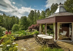 A covered patio with a table and chairs, surrounded by a garden with various plants and flowers. - Lodge for up to 10 in Rothiemurchus Forest with hot tub. (Aviemore)